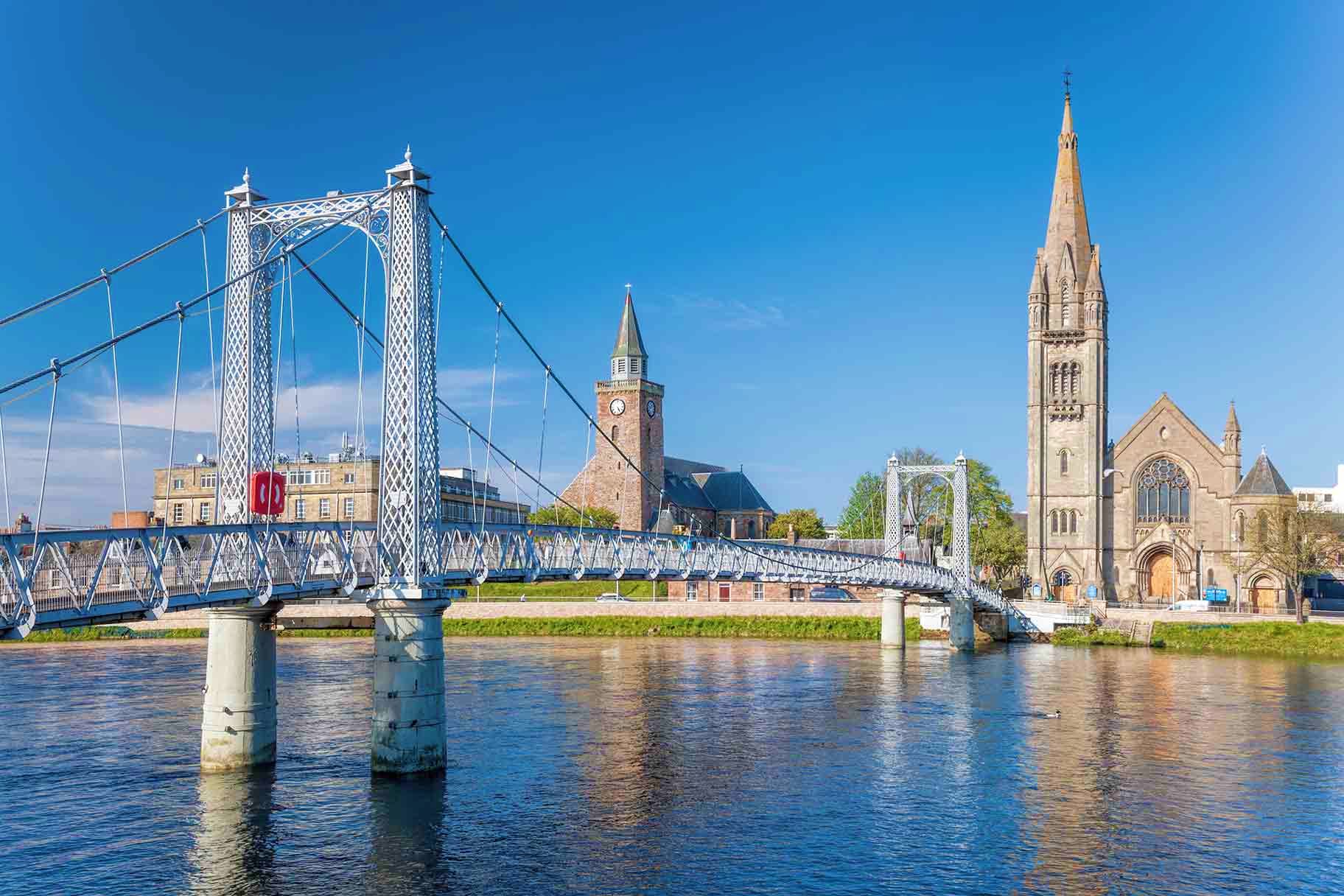 White suspension bridge spanning river with Gothic church spire and historic stone buildings under blue sky in Ireland